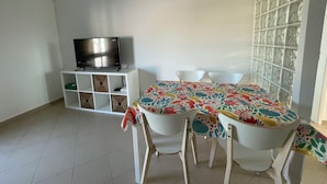 A dining area with a table covered by a floral tablecloth, surrounded by white chairs. A television is placed on a white cabinet with open shelves.