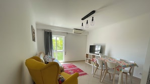 A living room with a yellow sofa, a dining table with chairs, a TV on a stand, and a window with curtains.