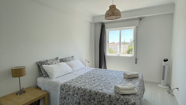 A bedroom with a bed, a bedside table with a lamp, a window with curtains, and a ceiling fan.