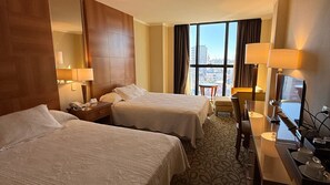 Une chambre d’hôtel avec deux lits, un bureau avec un ordinateur, une chaise et une grande fenêtre donnant sur la ville.