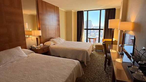 Ein Hotelzimmer mit zwei Betten, einem Schreibtisch mit Computer, einem Sessel und einem großen Fenster mit Blick auf die Stadt.