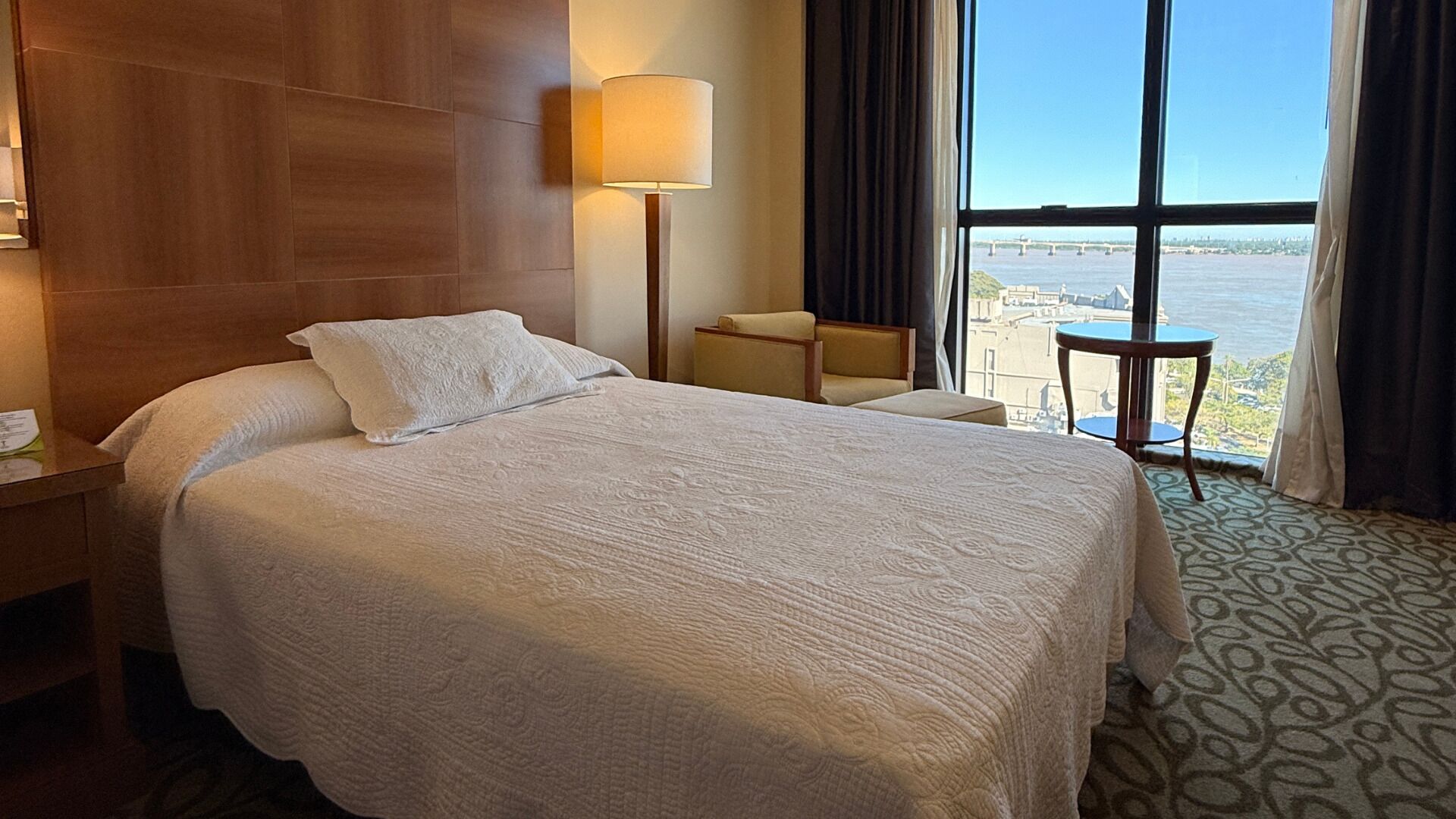 Habitación de hotel con una cama grande, una lámpara de noche, una silla, una mesita y vistas a un puente y al agua.