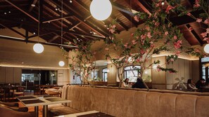 Een restaurant met hoge plafonds, zichtbare balken en hangende吊灯. Er zijn houten tafels en zitplaatsen met kussens.