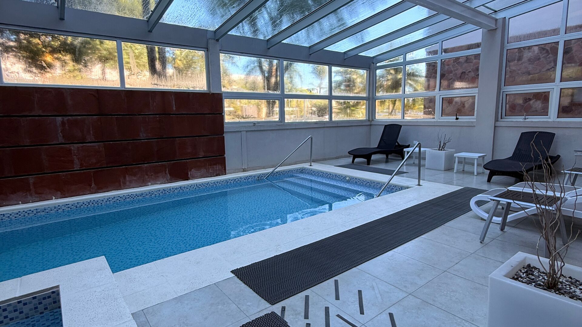 An indoor swimming pool with clear glass walls, a tiled floor, and a few lounge chairs.