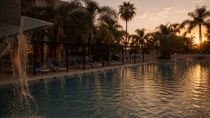 Piscina con sillas de descanso y palmeras, rodeada por un edificio con zona techada.
