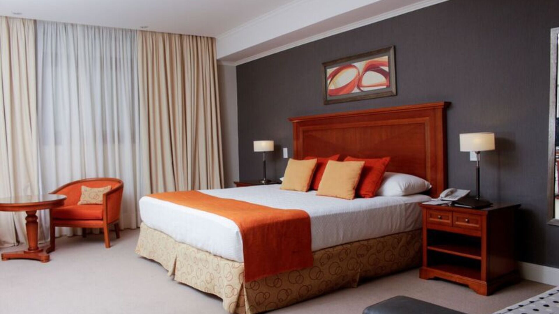 A hotel room with a large bed, a desk, a chair, a nightstand, and a painting on the wall.