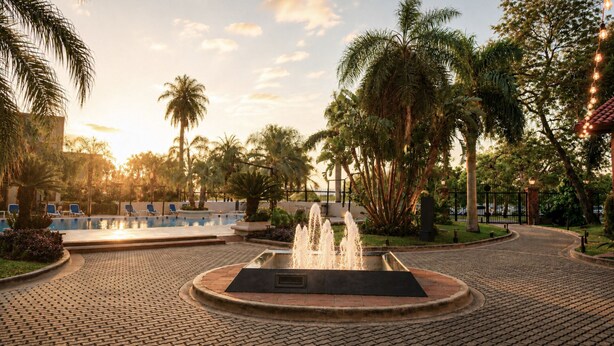 Zona de piscina con fuente, palmeras y un camino pavimentado.