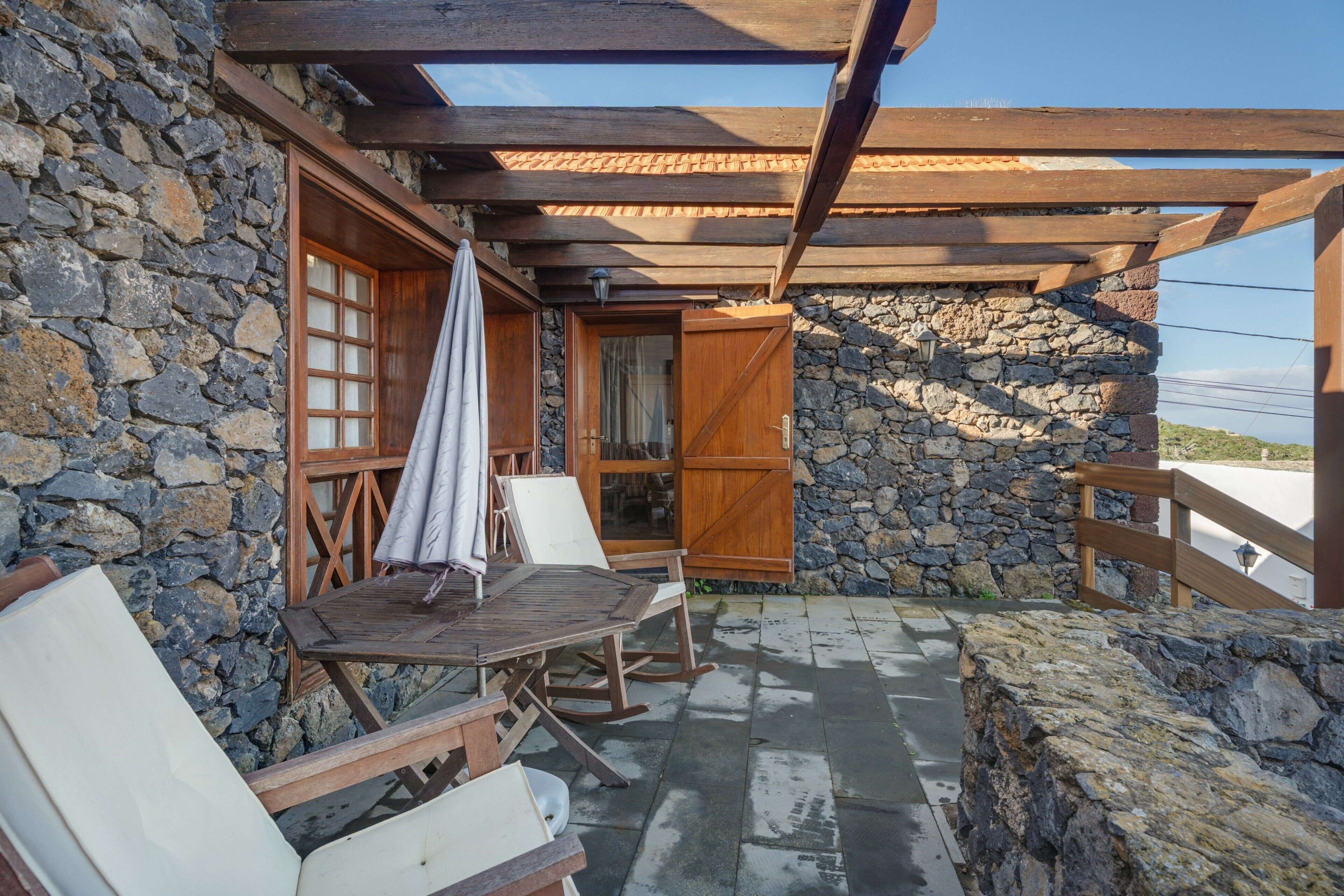 A stone building with a wooden pergola, a table with chairs, and a view of the landscape.