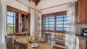 Un comedor con mesa de madera, sillas, un cuenco de fruta, tazas y una cafetera. Hay ventanas grandes con cortinas y vistas al exterior.