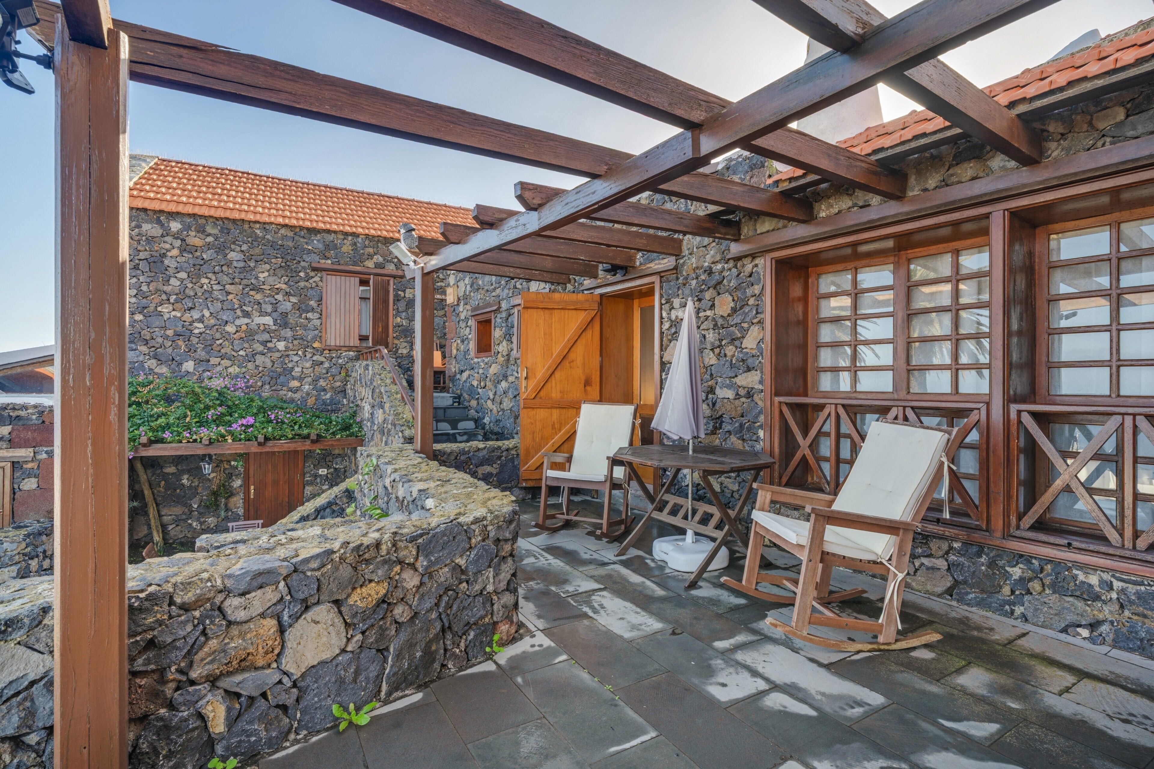 Un edificio de piedra con una pérgola de madera, un muro de piedra y una mesa de madera con sillas.