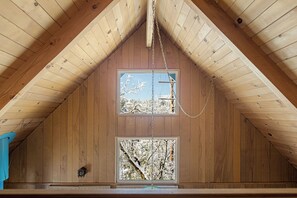 A room with wooden walls and a sloped ceiling, featuring two windows with views of trees and a clear sky.