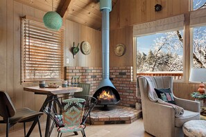 A cozy room with a fireplace, wooden walls, and a view of snow-covered trees. - MCM Vintage Fireplace•Views•Fully Fenced•BBQ•Large Deck (Lake Arrowhead)