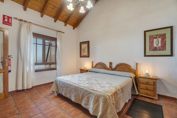 Une chambre avec un plafond en bois, un lit recouvert d’une couvre-lit à motifs, une table de chevet avec une lampe, un tableau encadré au mur et une fenêtre avec des rideaux.