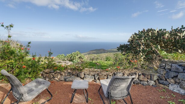 A patio with wicker chairs and a stone wall, overlooking a coastal landscape with cacti and a view of the ocean. - Country House 'El Almendro' with Sea Views, Shared Terrace and Wi-Fi (El Mocanal)