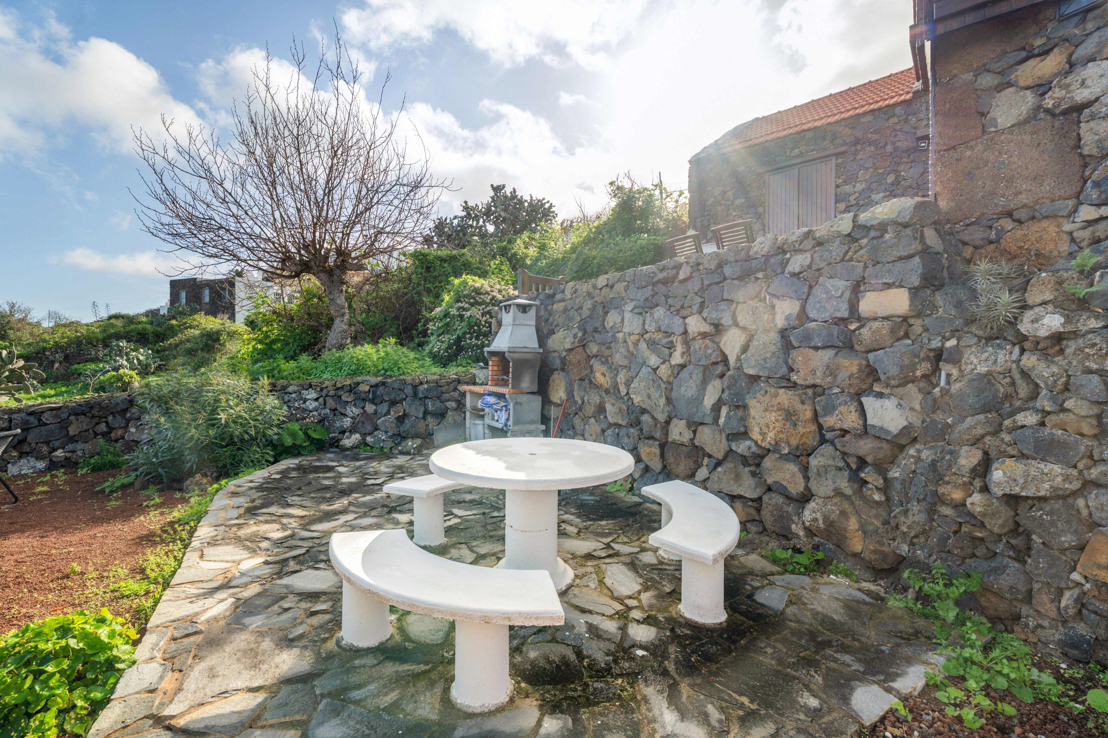 Un mur de pierre agrémenté d’un banc et d’une table en pierre, le tout entouré de verdure et sous un ciel dégagé.