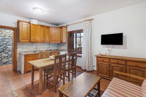 A kitchen with wooden cabinets, a dining table, chairs, a TV mounted on the wall, and a stone wall. - Country House 'El Almendro' with Sea Views, Shared Terrace and Wi-Fi (El Mocanal)