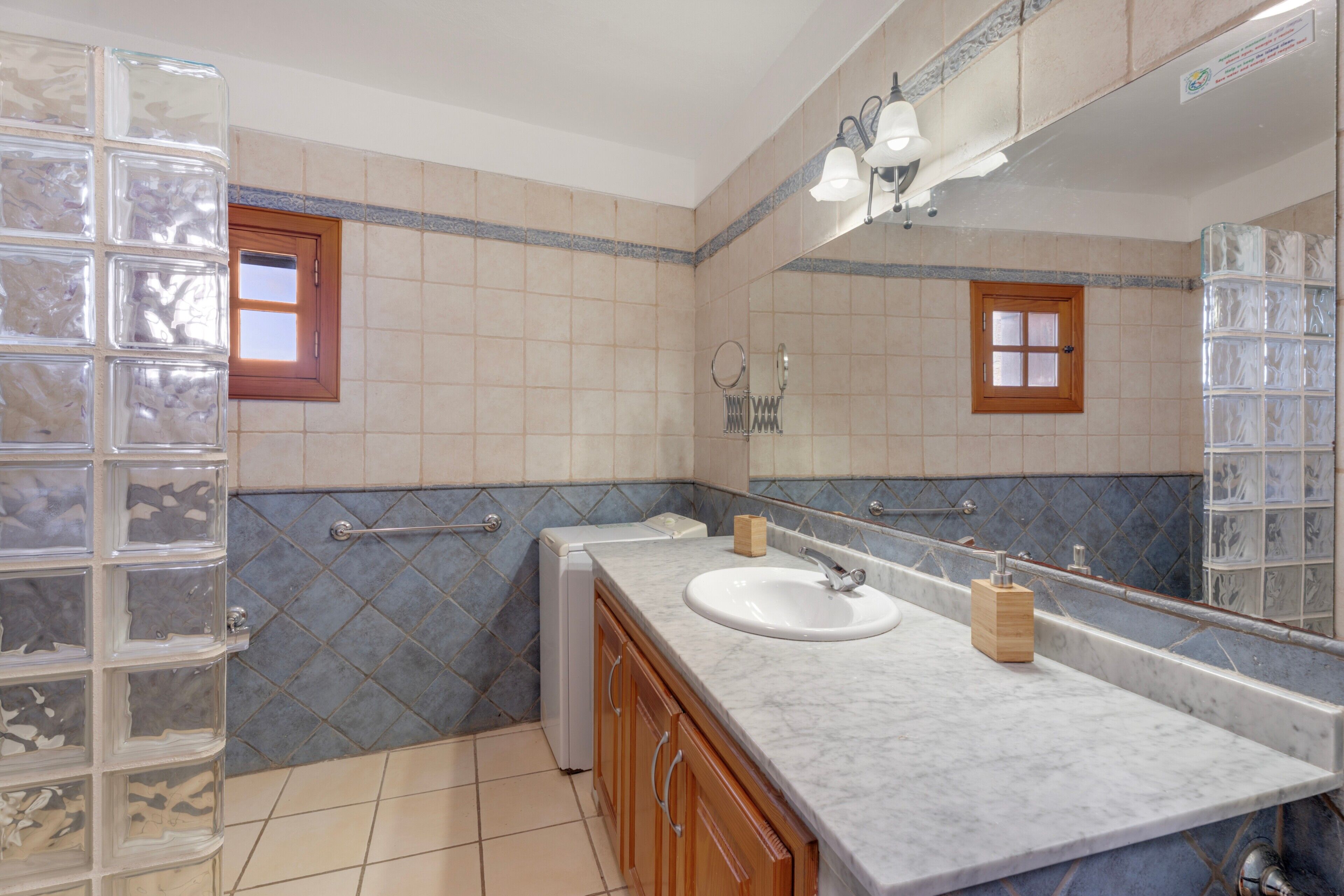 Un baño con lavamanos blanco, armarios de madera, cabina de ducha de vidrio y pared con azulejos y un ventanal.