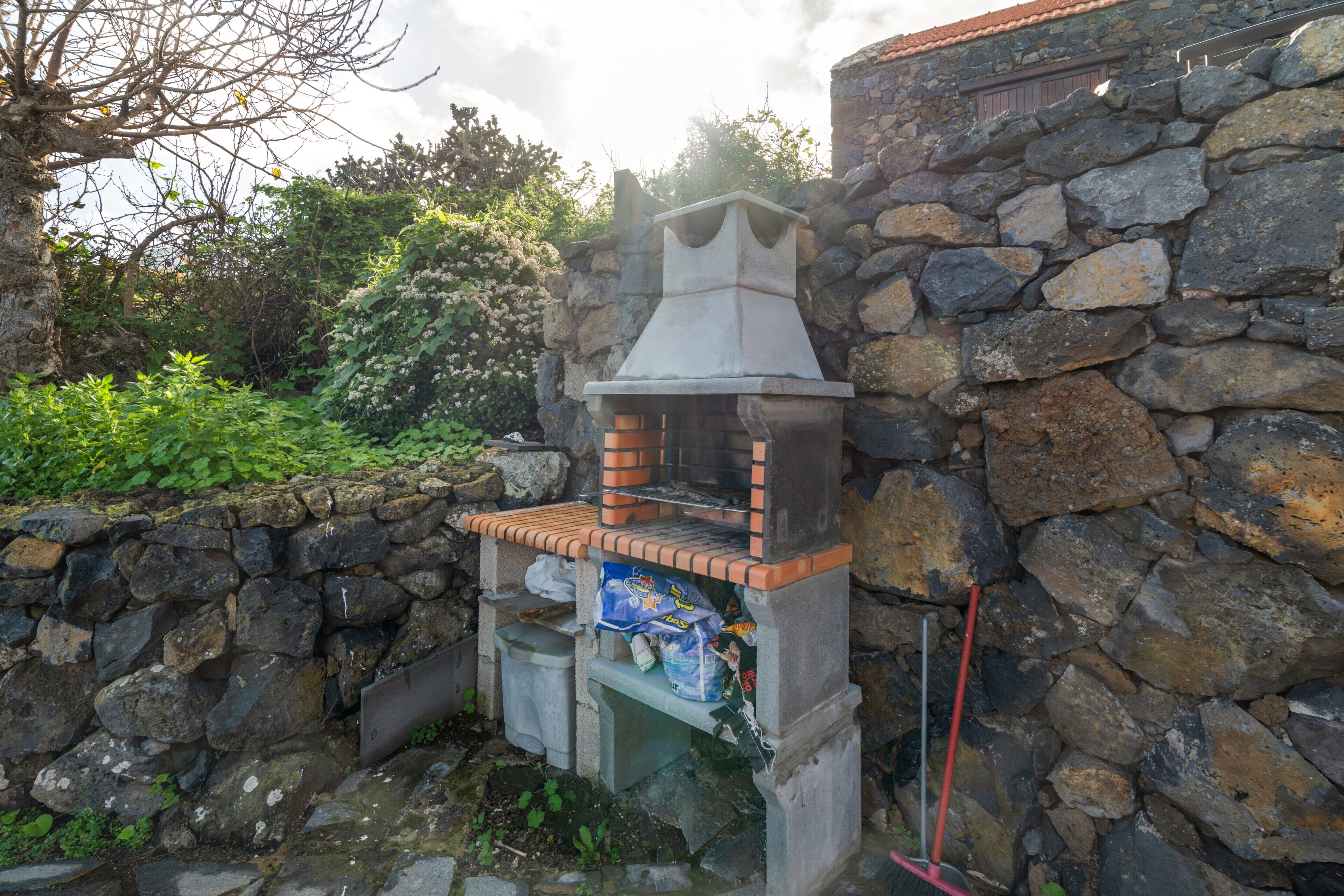 A stone-built outdoor structure with a chimney, possibly a barbecue or grill area, surrounded by greenery and a stone wall.