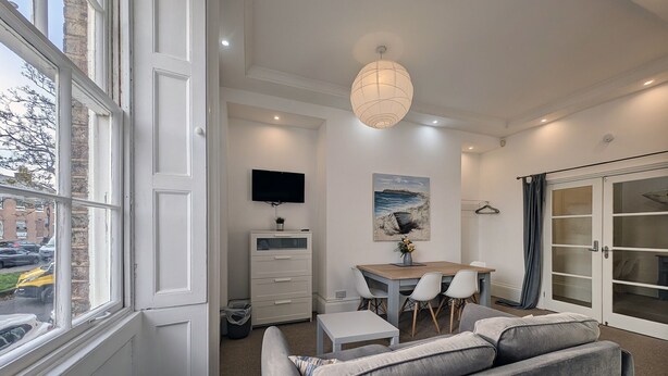 A modern living room with a sofa, a dining table with chairs, a TV mounted on the wall, and a large window. - Pass the Keys | Coastal 2 - Bedroom Flat in Tynemouth | Sleeps 5 (North Shields)