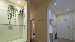 A hallway with a glass shelf holding books and a hanging plant, leading to a door with a small bench and a wall-mounted artwork. - Pass the Keys | Coastal 2 - Bedroom Flat in Tynemouth | Sleeps 5 (North Shields)