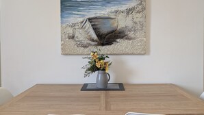A dining table with a vase of flowers and a painting of a coastal scene. - Pass the Keys | Coastal 2 - Bedroom Flat in Tynemouth | Sleeps 5 (North Shields)