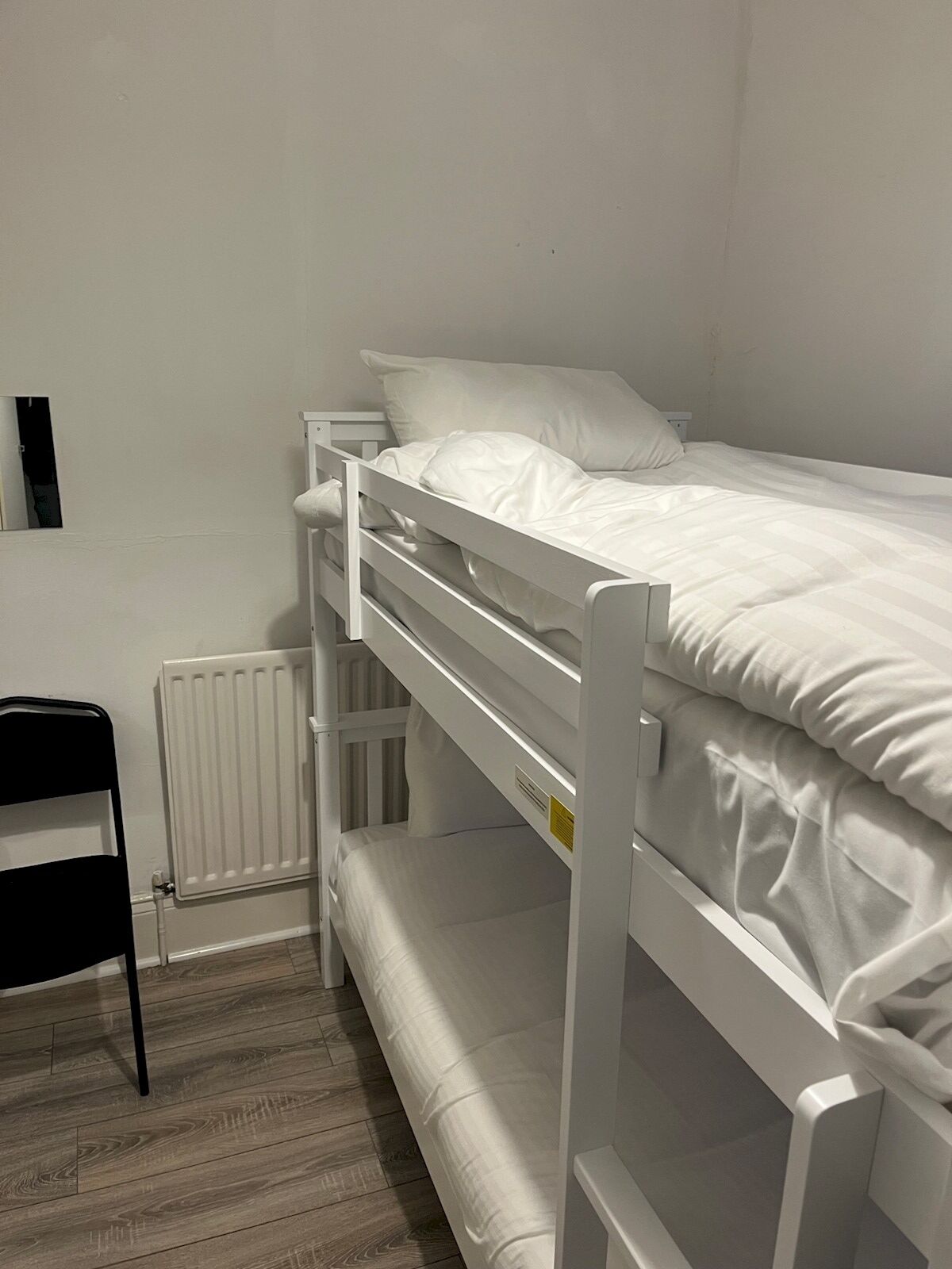 Een stapelbed met witte beddengoed, een raam met jaloezieën en een houten vloer.