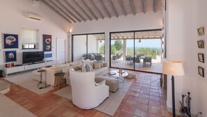 A spacious living room with a white sofa set, a flat-screen TV, and a large sliding glass door leading to an outdoor patio with a dining area. - Casa Velha: Pool & Ocean View (Loulé)