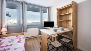 A compact hotel room with a bed, a desk with a chair, a TV, and a wooden shelf unit.