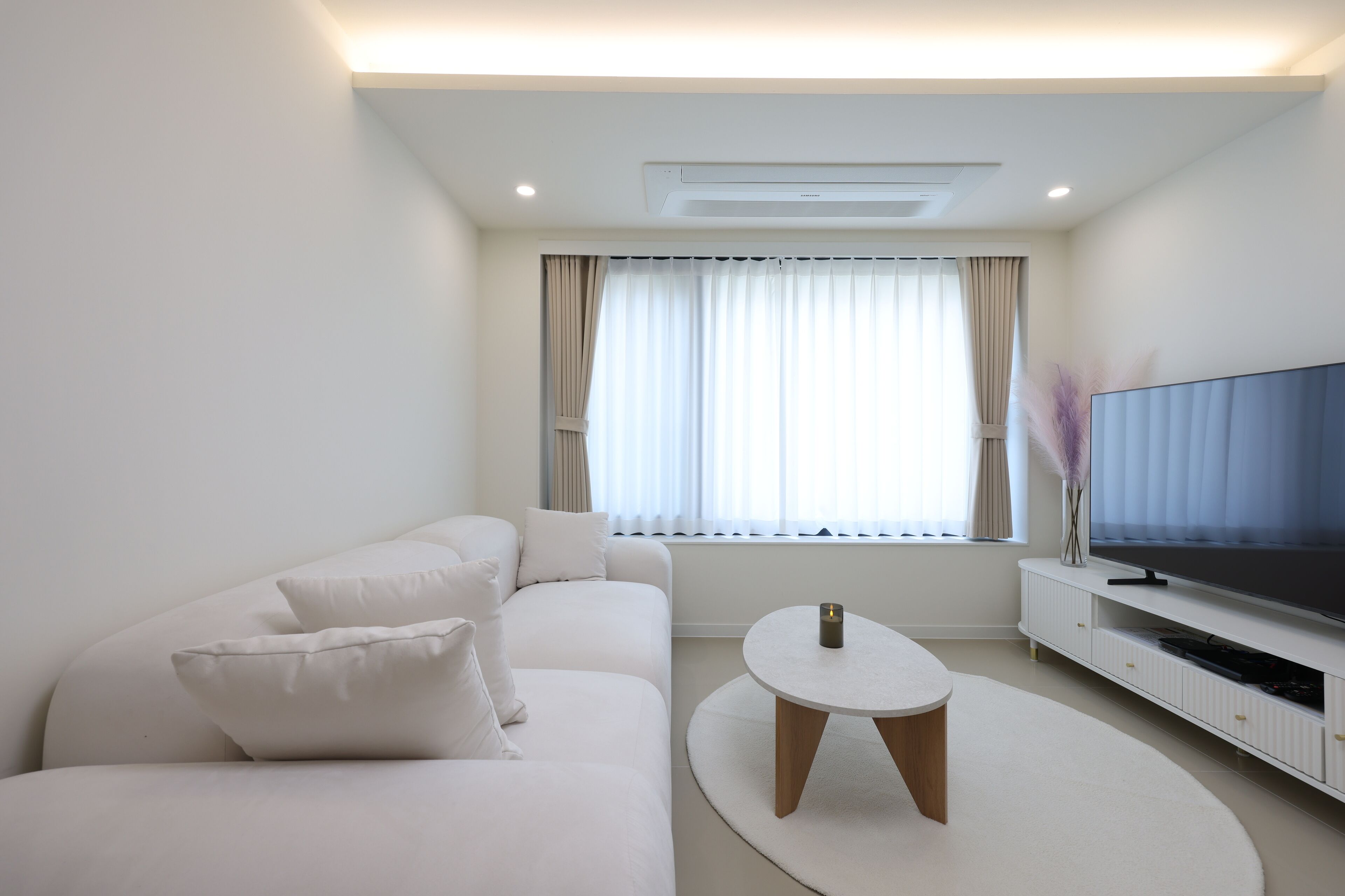 A modern living room with a white sofa, a round coffee table, a flat-screen TV, and a window with curtains.