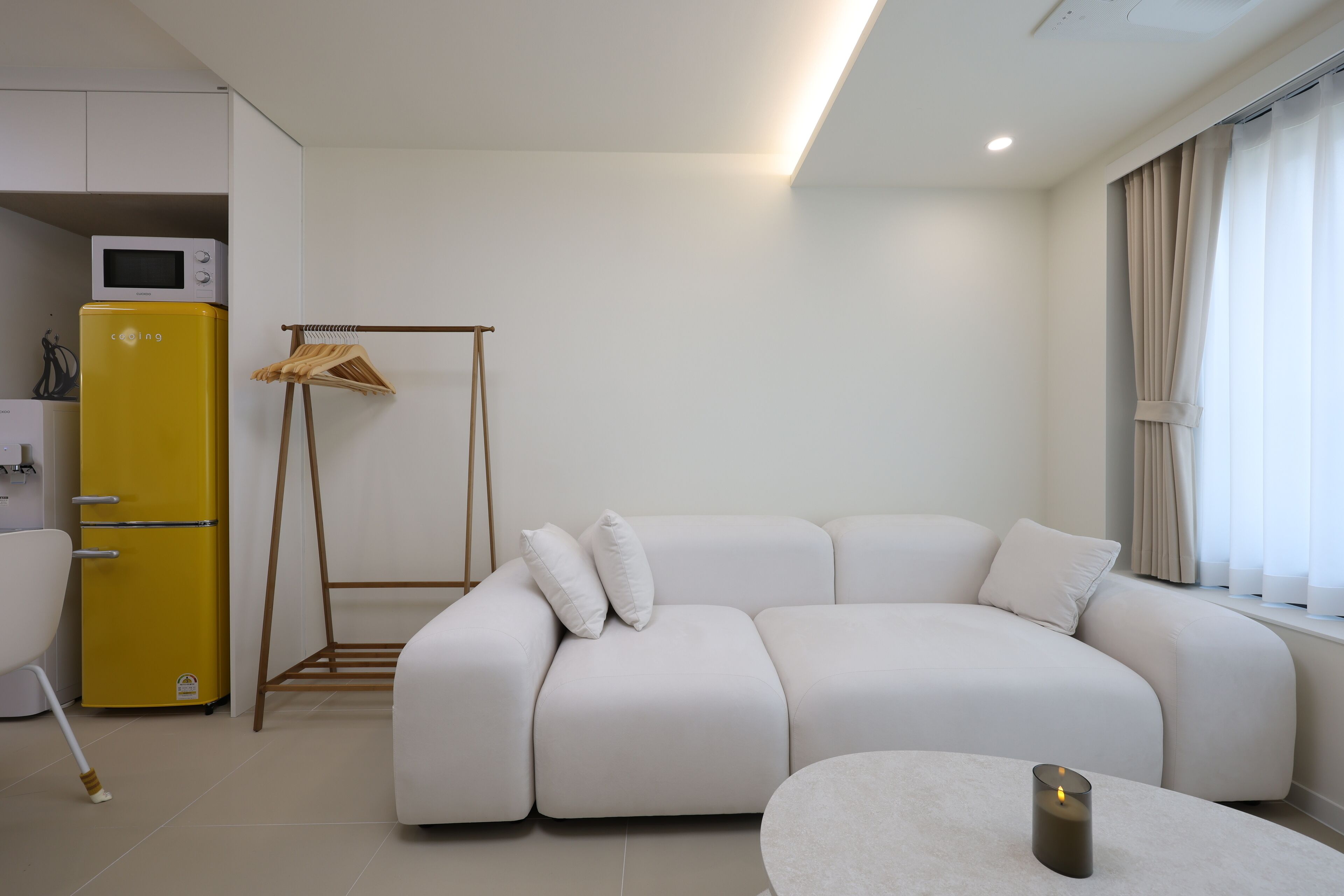 A modern living room with a white sectional sofa, a yellow mini-fridge, a wooden drying rack, and a small table with a candle.