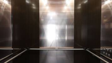 An elevator interior with metallic walls, a reflective floor, and overhead lighting.