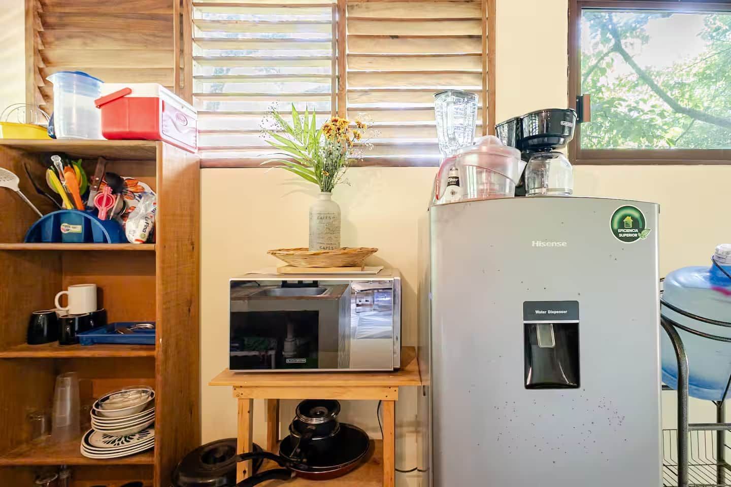 Eine Küchenzeile mit Kühlschrank, Mikrowelle und Wasserspender. Ein kleiner Tisch steht dort, auf dem sich eine Blumenvase befindet, und ein Fenster mit Jalousien ist zu sehen.