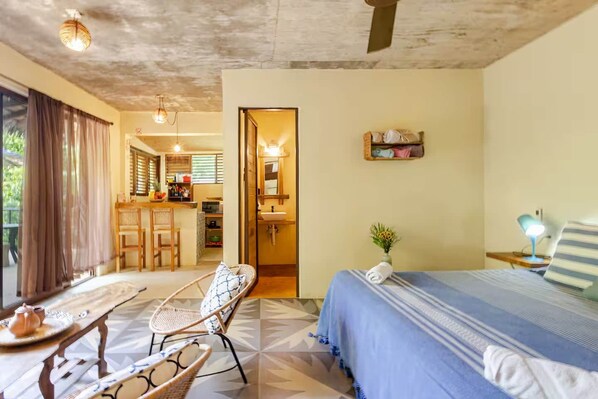 A hotel room with a bed, a chair, a small table, a desk, and a kitchenette.