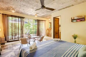 A hotel room with a large bed, a dining table, chairs, and a view of the outdoors through large windows.