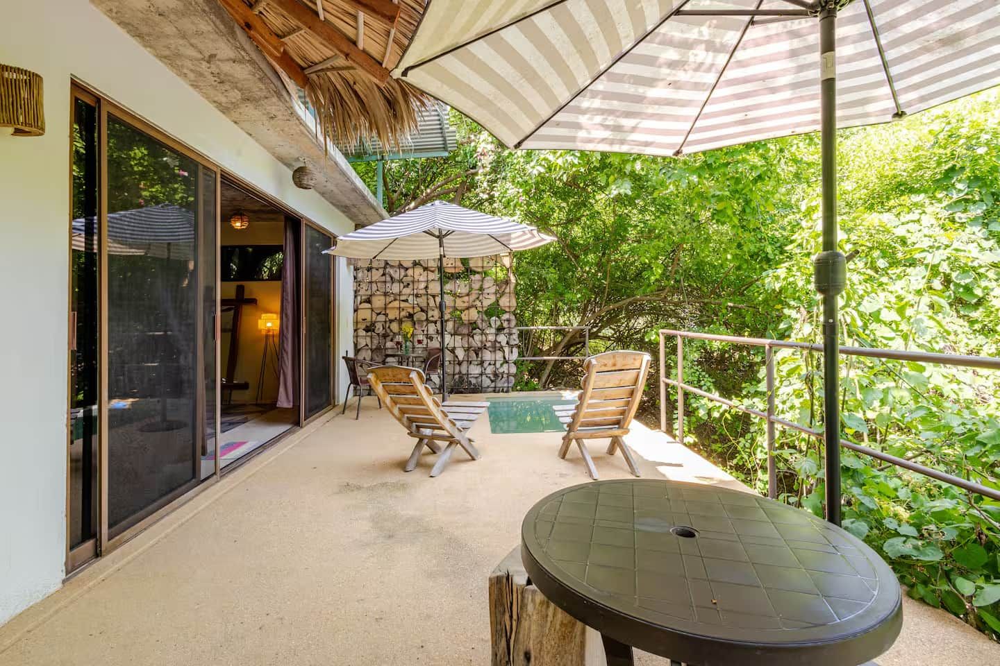 Un espacio al aire libre techado con piscina, dos sillas de madera, una mesa y un paraguas a rayas.