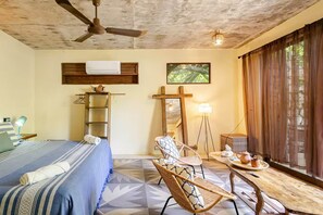 Ein Hotelzimmer mit einem Bett, einem Sessel, einem Tisch mit Tablett, einer Stehlampe, einem Deckenventilator und einem großen Fenster mit Vorhängen.