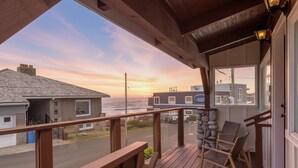 Une terrasse en bois, agrémentée de chaises en osier, donnant sur une rue résidentielle et offrant une vue sur l’océan au coucher du soleil.