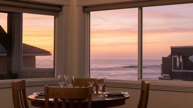 Une table à manger avec des chaises et des verres à vin, offrant une vue sur le coucher de soleil et l’océan.