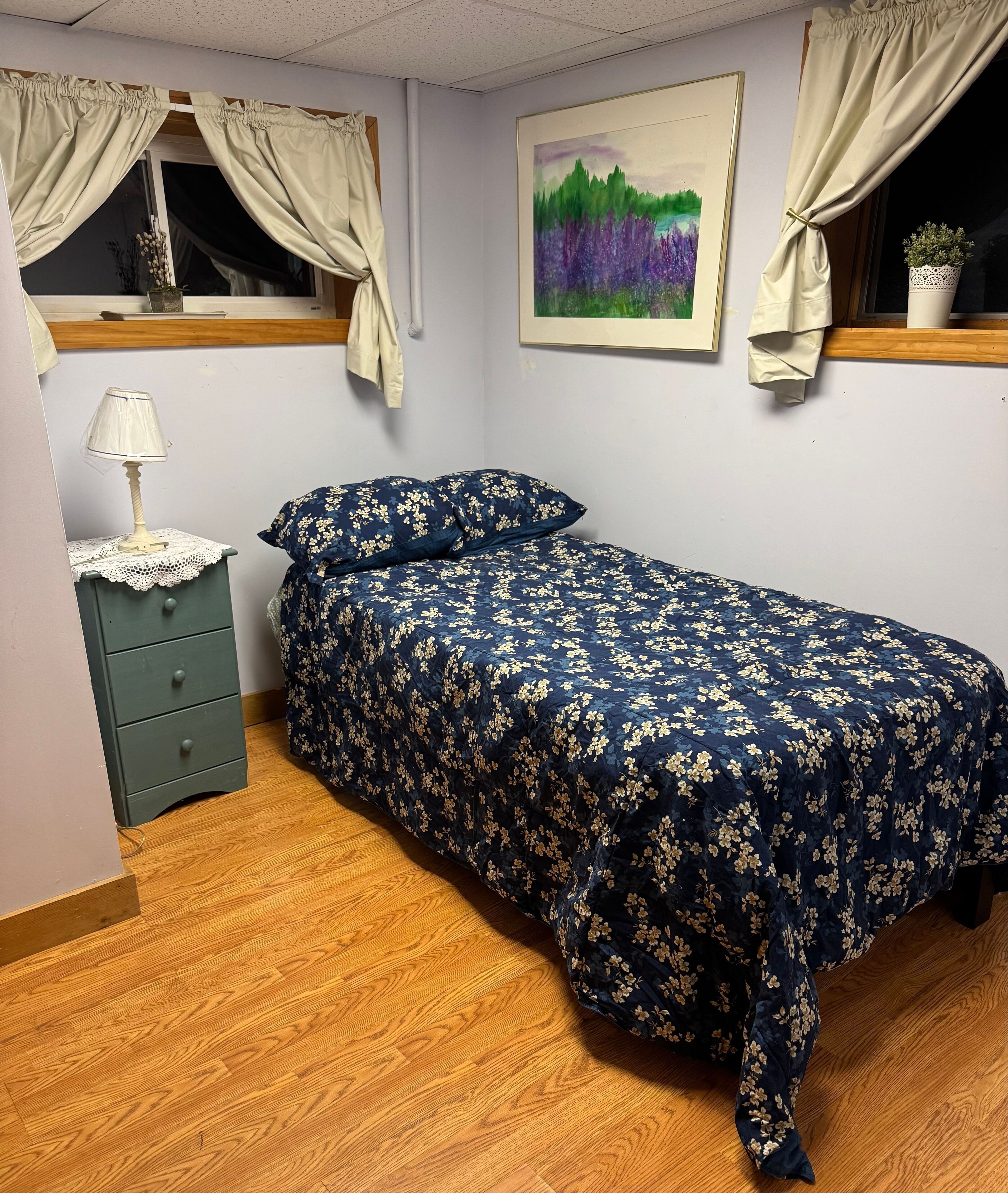 A bedroom with a bed, a nightstand, a lamp, a window with curtains, a framed painting, and a potted plant.