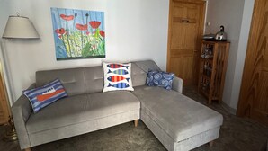 Un divano angolare grigio con tre cuscini, una lampada, un quadro e un mobiletto in legno.