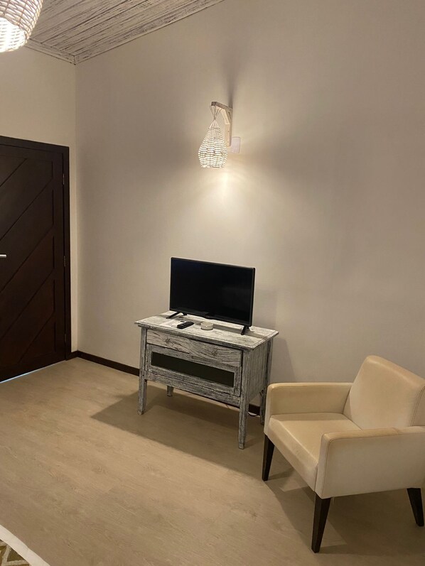 Une pièce avec une porte en bois foncé, un canapé blanc, un meuble TV en bois sur lequel est posée une télévision à écran plat, et un luminaire fixé au mur.