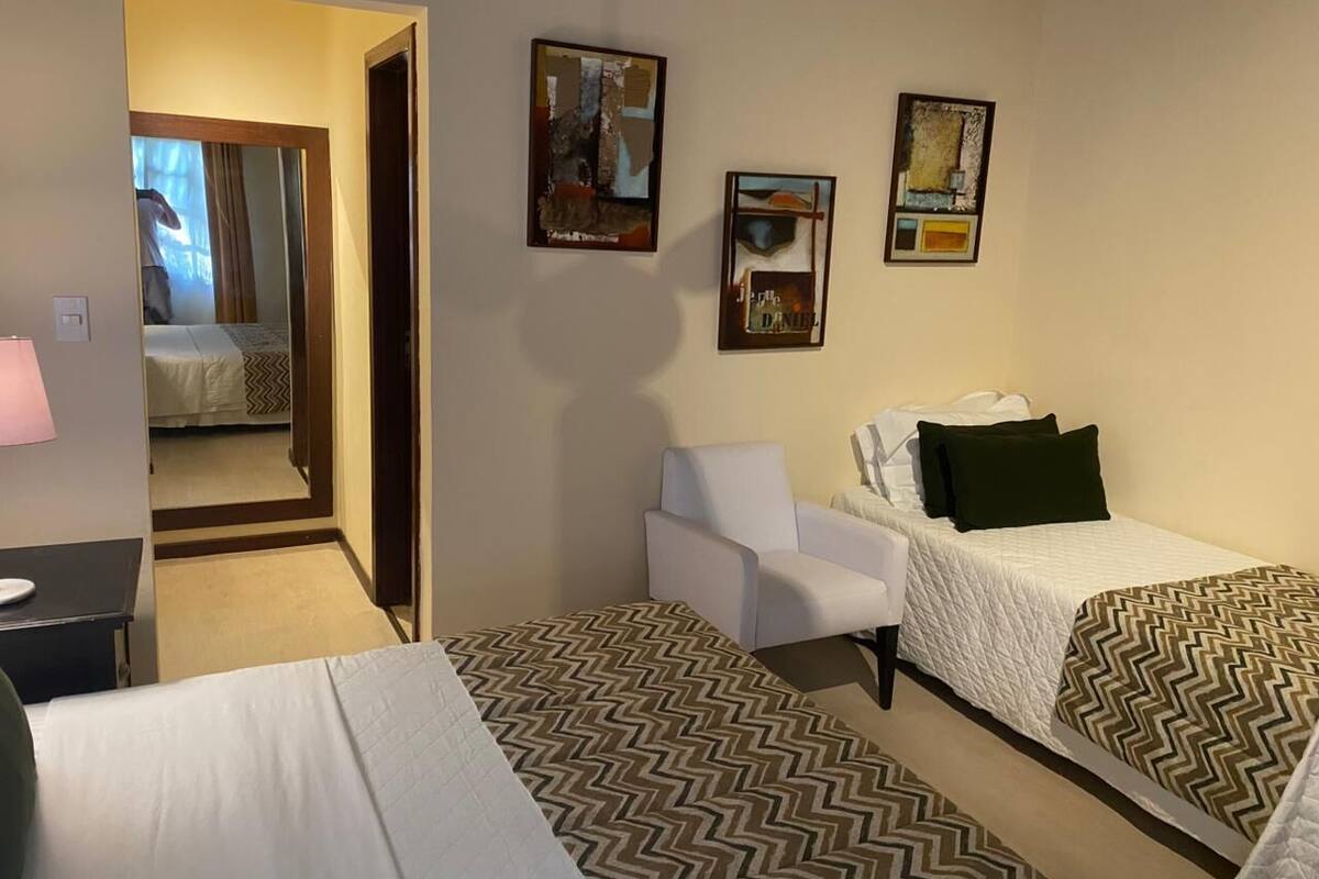 Ein Hotelzimmer mit zwei Einzelbetten, einem Sessel, einem Schreibtisch und einem Spiegel. An der Wand hängen zwei Bilder.