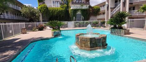 A swimming pool with a fountain, surrounded by a white fence and a building with ivy-covered walls.