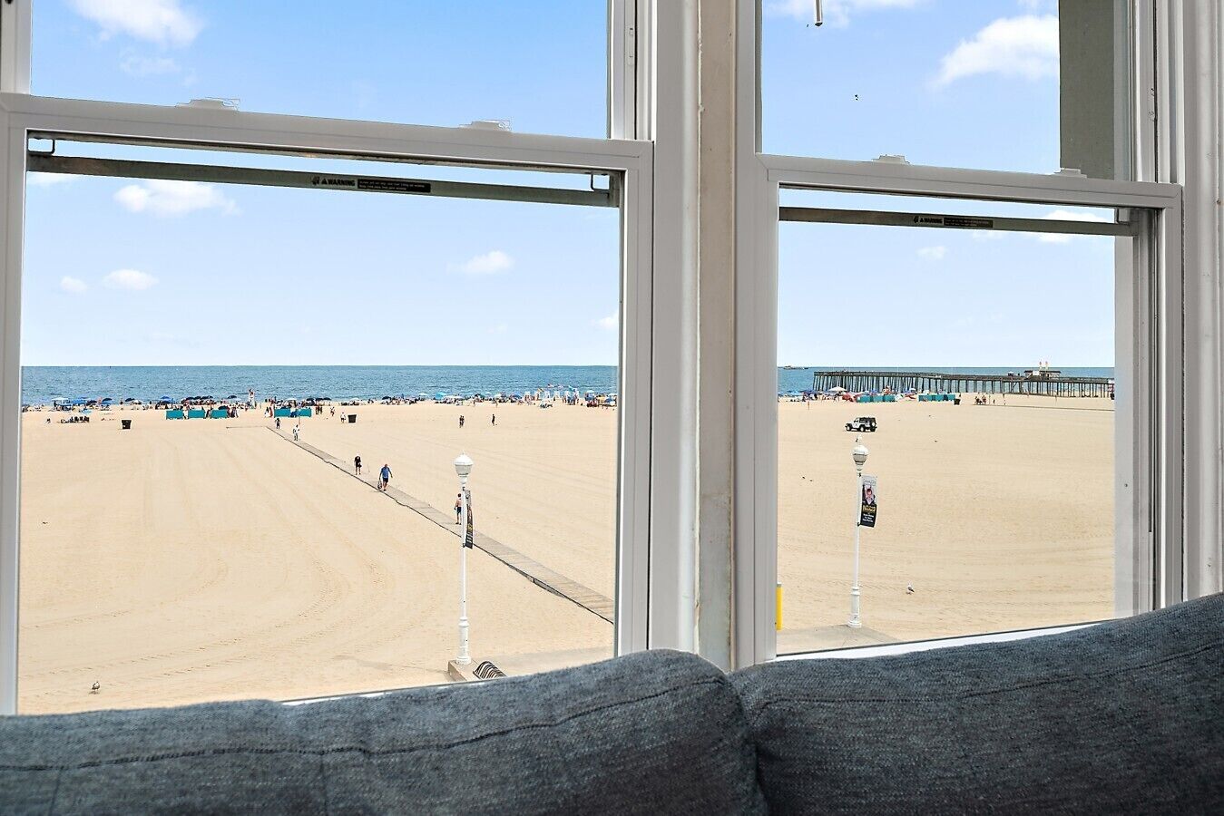A beach view through a window with a white frame, a sandy beach, a pier, and a clear sky.
