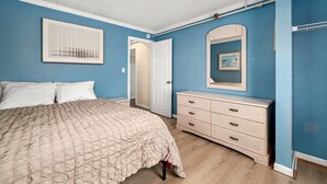 Un dormitorio con cama, cómoda, puerta y espejo adosado a la pared.