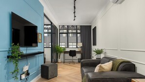 A modern living room with a flat-screen TV, a grey sofa, a wooden coffee table, and a desk with a chair.