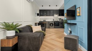 A modern living room with a grey sofa, a small wooden side table, and a potted plant. A kitchen with grey cabinets and a built-in oven is visible in the background.