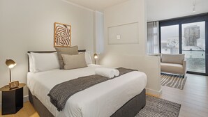 Quarto de hotel moderno com uma cama grande, mesa de cabeceira com abajur, vista da cidade por grandes janelas e varanda com sofá.