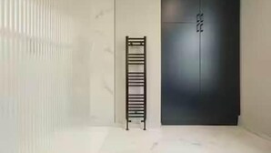 Een kamer met een hoge, smalle, zwarte metalen radiator, een donkere garderobe en een lichtgekleurde betegelde vloer.