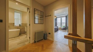 Ein Flur mit Holzboden, der zu einem Badezimmer mit Badewanne und Toilette führt. Ein Heizkörper steht auf dem Boden und ein gerahmtes Kunstwerk ist an der Wand angebracht.